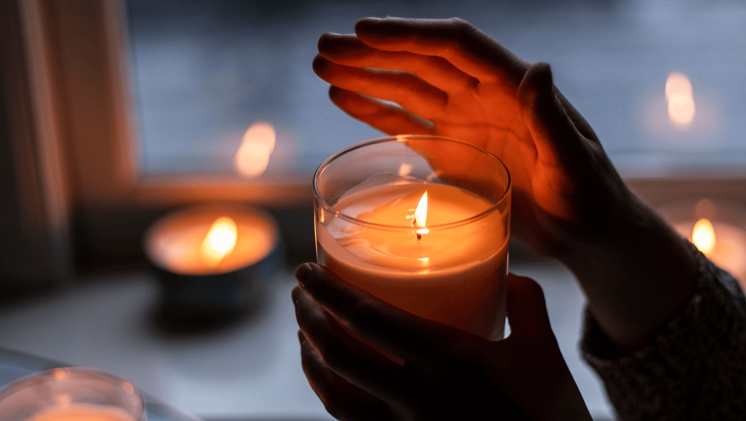 Nainen pitää kädessään kynttilää  ja suojaa sen liekkiä toisella kädellään, kun taustalla on hämärää ja ikkunalaudalla palaa useita kynttilöitä