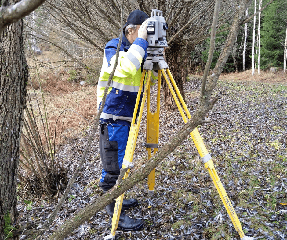 Henkilö työasussa kurkistaa kolmijalan päällä seisovan mittalaitteen läpi.