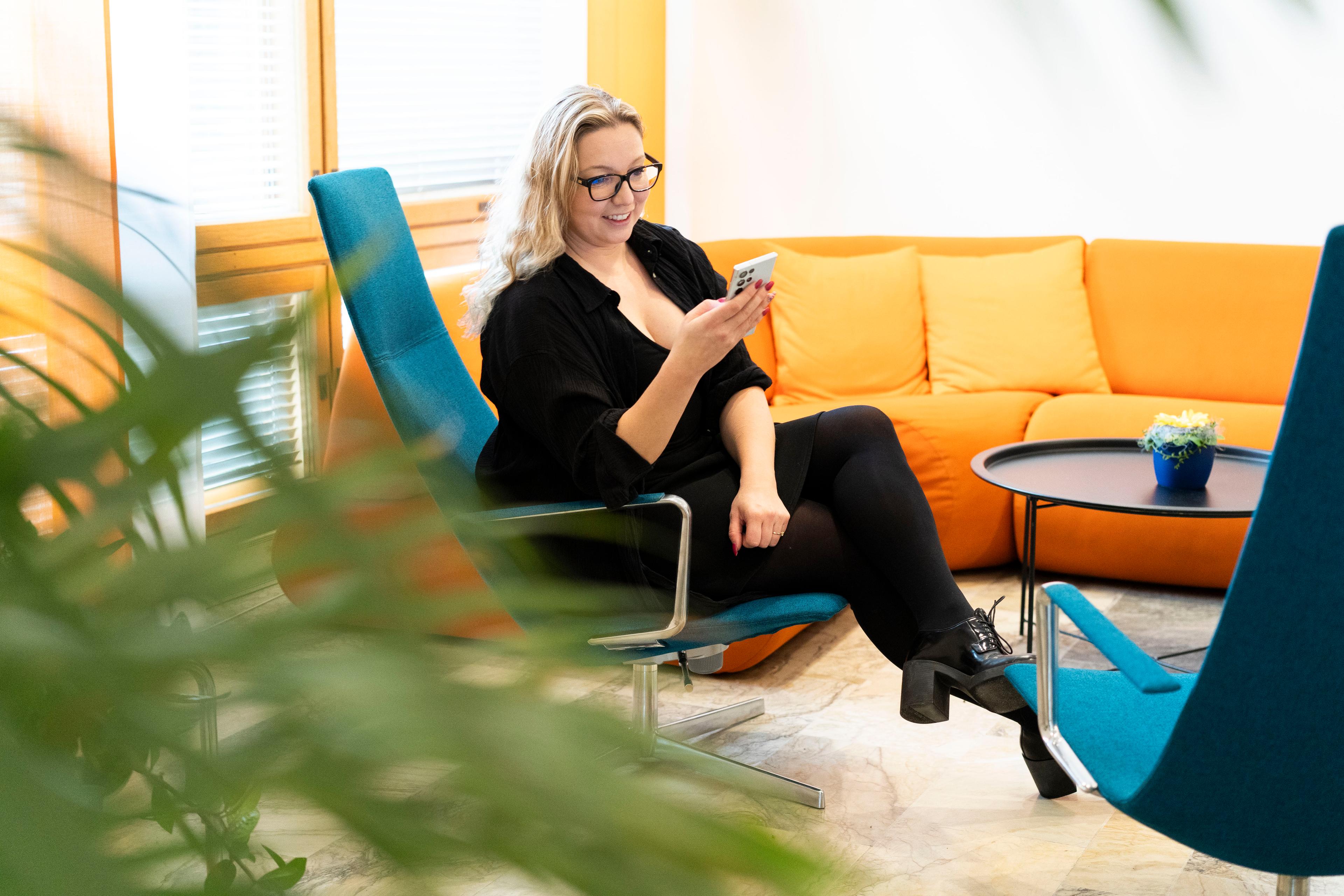 Mustiin vaatteisiin pukeutunut vaaleahiuksinen nainen istuu sinisellä tuolilla ja katsoo hymyillen puhelintaan. Etualalla on viherkasvi, takana näkyy oranssi kulmasohva ja pieni sohvapöytä. Kuvan tunnelma on raikas.