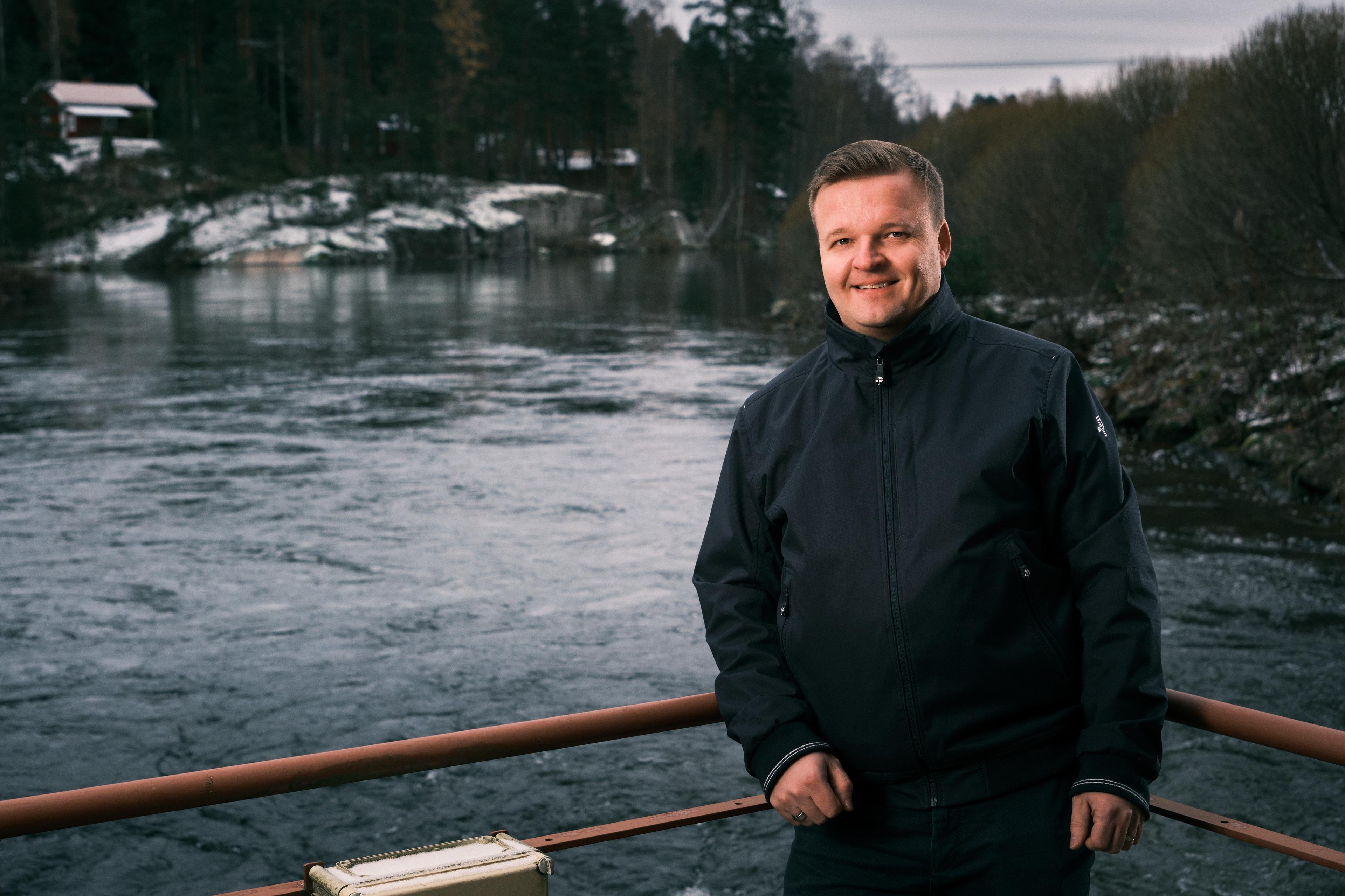 Verlan vesivoimalan edustalla alkutalvesta otettu kuva, jossa mustatakkinen mies katsoo suoraan kameraan ja hymyilee. Mies nojaa ruskeaan kaiteeseen ja alhaalla on virtaava vesi. Takana näkyvällä vastarannalla on pieni talo, puustoa ja kalliorantaa.