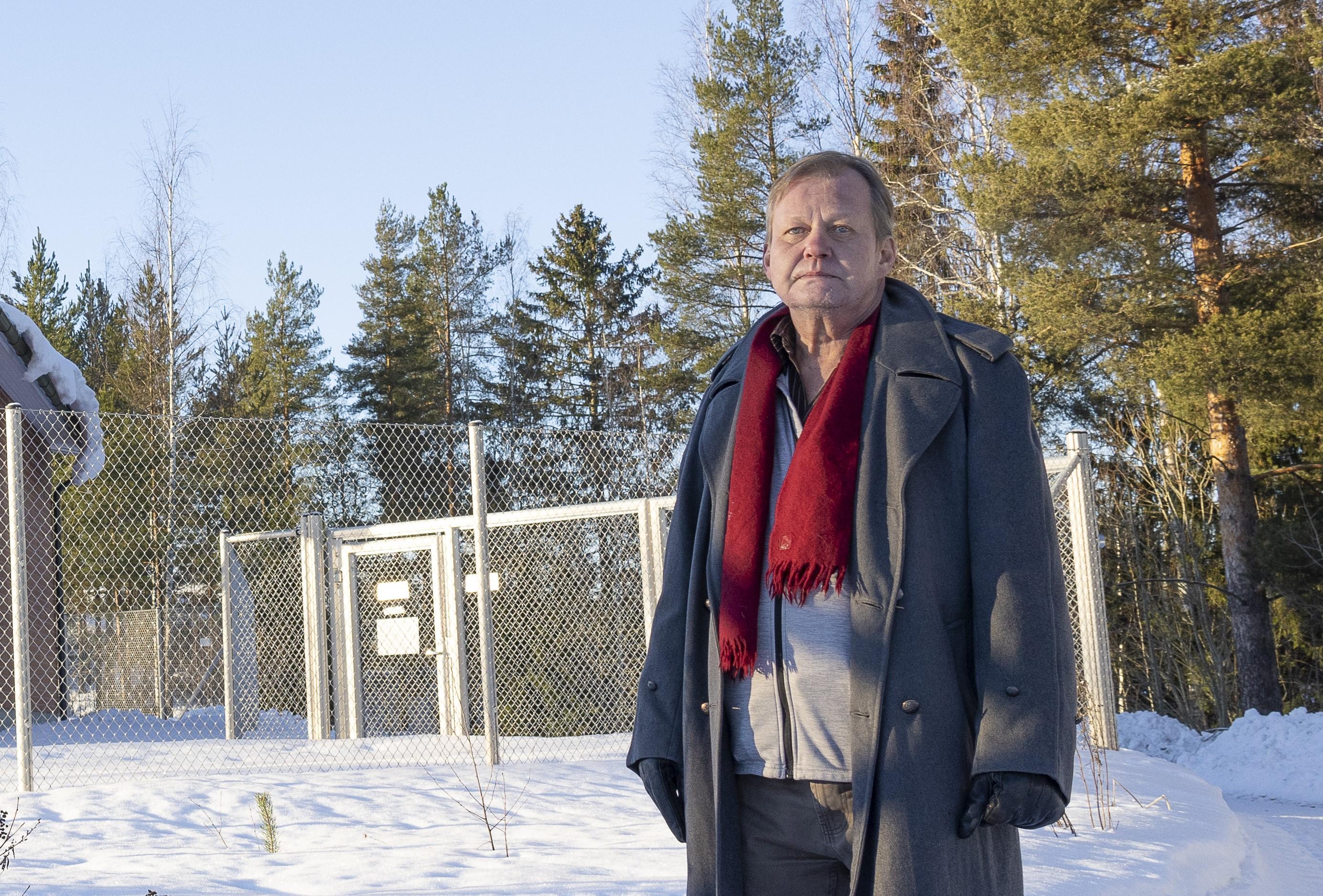 Avoinna olevaan harmaaseen talvitakkiin ja punaiseen kaulaliinaan pukeutunut mies seisoo mietteliäänä talvisessa maisemassa. Mies katsoo hieman ohi kameran linssin. Taustalla näkyy harmaalla verkkoaidalla aidattu tila. Aidassa on lukittu ovi. Kuvan vasemmassa reunassa pilkistää punatiilinen rakennus. Miehen ja aidan takana on havumetsää.