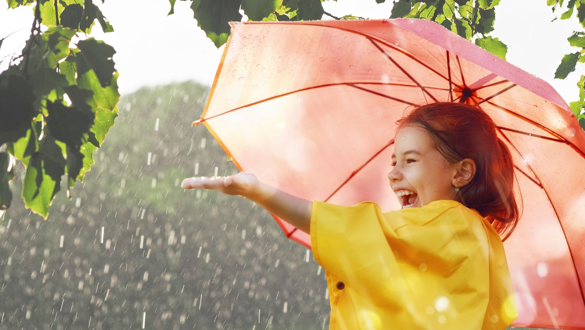 Pieni ruskeahiuksinen tyttö keltaisessa sadetakissa ja vaaleanpunaisen sateenvarjon alla ojentaa vasemman käden kohti sadepisaroita. Taustalla näkyy vihreää ja auringon pilkahduksia.