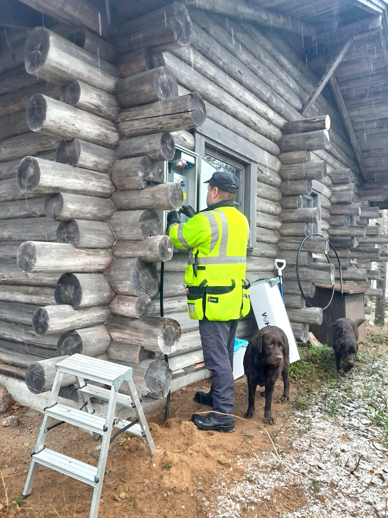 Keltaisiin työmaavaatteisiin pukeutunut mies tekee sähköasennusta harmaan hirsimökin ulkoseinustalla. Miehellä on siniharmaa hattu ja samanväriset housut, hän on selin kameraan. Asentajan vieressä näkyy kaksi ruskeaa koiraa sekä harmaa työjakkara. Kuvan oikeassa laidassa näkyvällä nurmella on kevyt lumipeite.