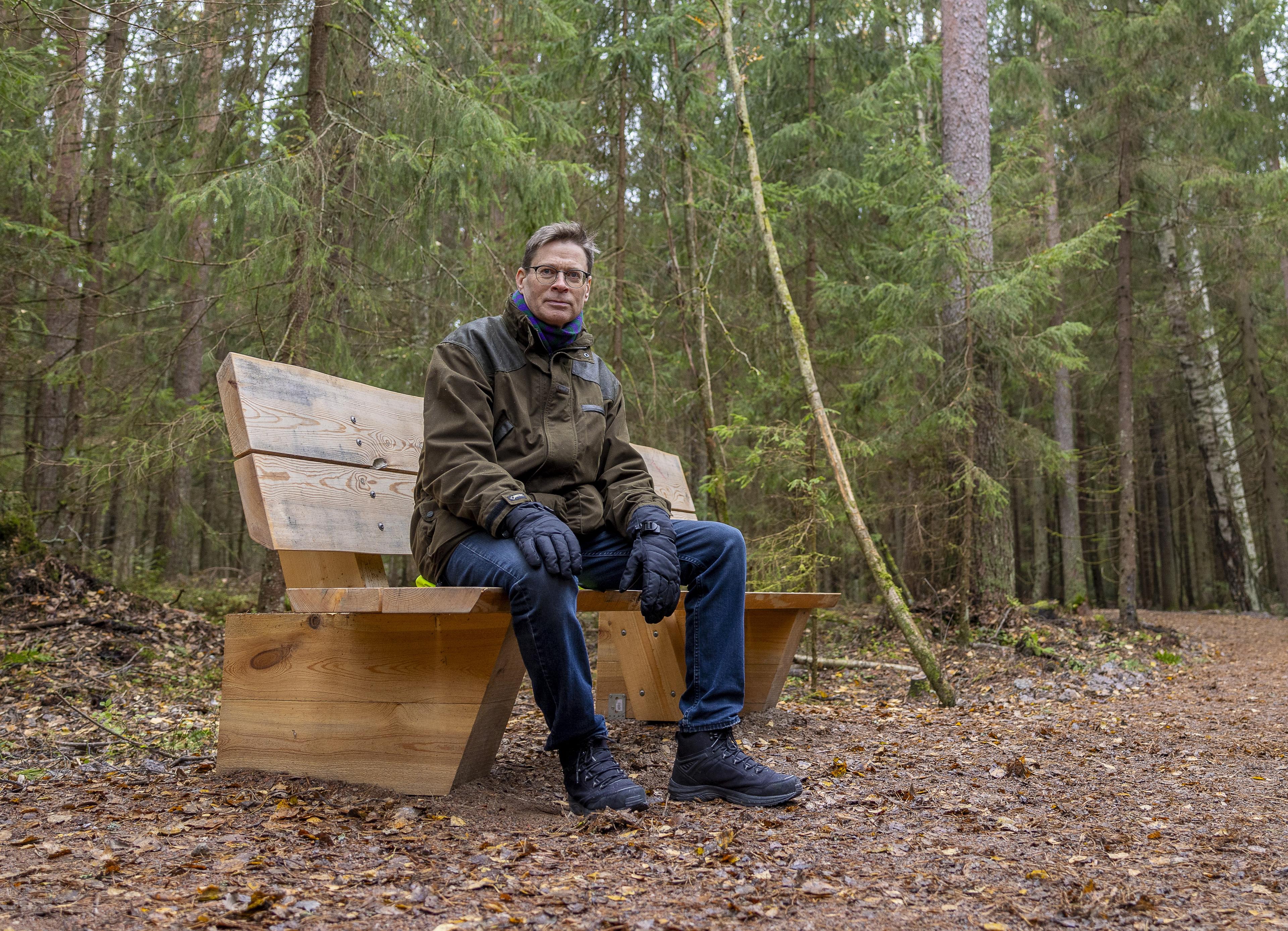 Keski-ikäinen silmälasipäinen mies istuu metsätien varrella vaalealla puupenkillä. Taustalla näkyy kuusimetsää ja muutama koivu, vuodenaika kuvassa on loppusyksy.