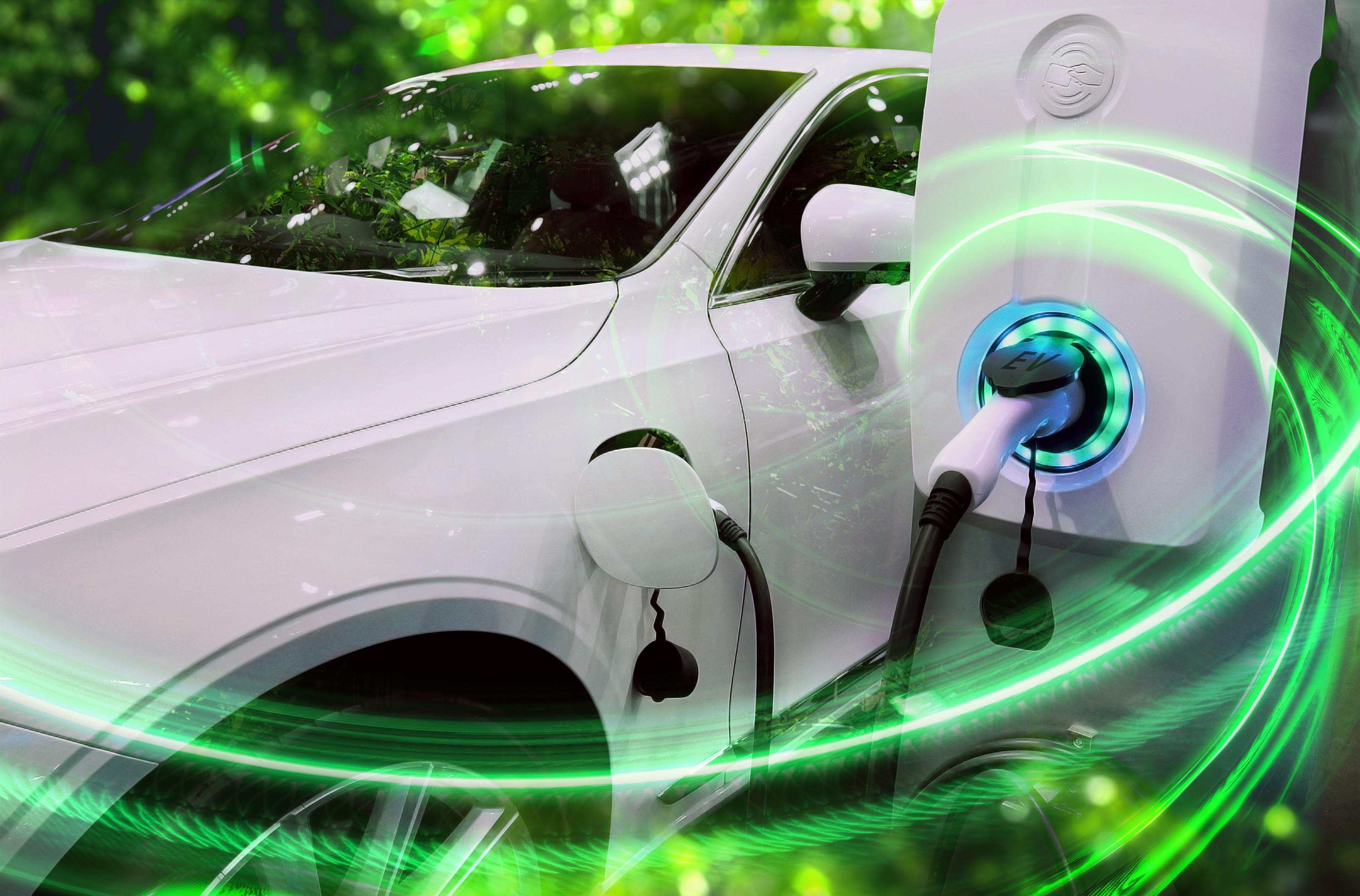 Kuvassa näkyy valkoinen tummalasinen sähköauto latauksessa sekä valkoinen sähköautojen latauslaite, johon latausjohto yhdistyy. Kuvan taustalla näkyy vihreää valoa ja etuosassa on vihreitä valojuovia.