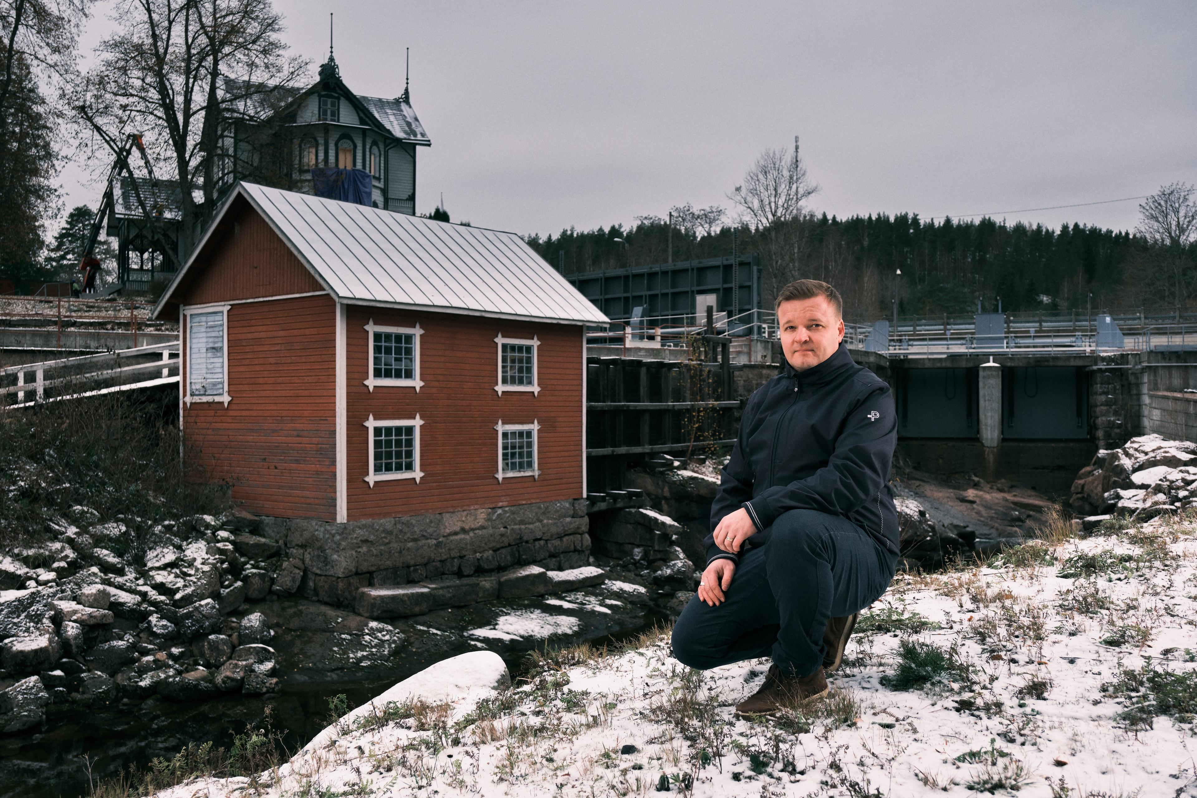 Mustissa talvivaatteissa oleva mies on kyykistynyt kevyen lumipeitteen peittämään maahan, katse suoraan kameraan. Taustalla on vesivoimalaitos, punainen puutalo sekä taustalla vanhempi kartanorakennus.