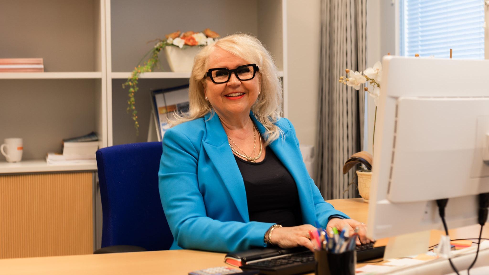 Sinijakkuinen ja vaaleahiuksinen nainen istuu toimistossa työpöydän äärellä ja katsoo hymyillen suoraan kameraan. 