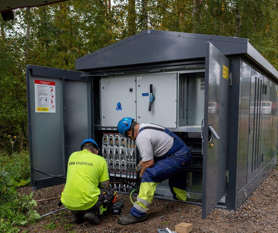 Metallisen harmaan puistomuuntamon ovet auki ja kaksi henkilöä suojavarusteissa kytkemässä kaapeleita kytkimiin sen sisälle.