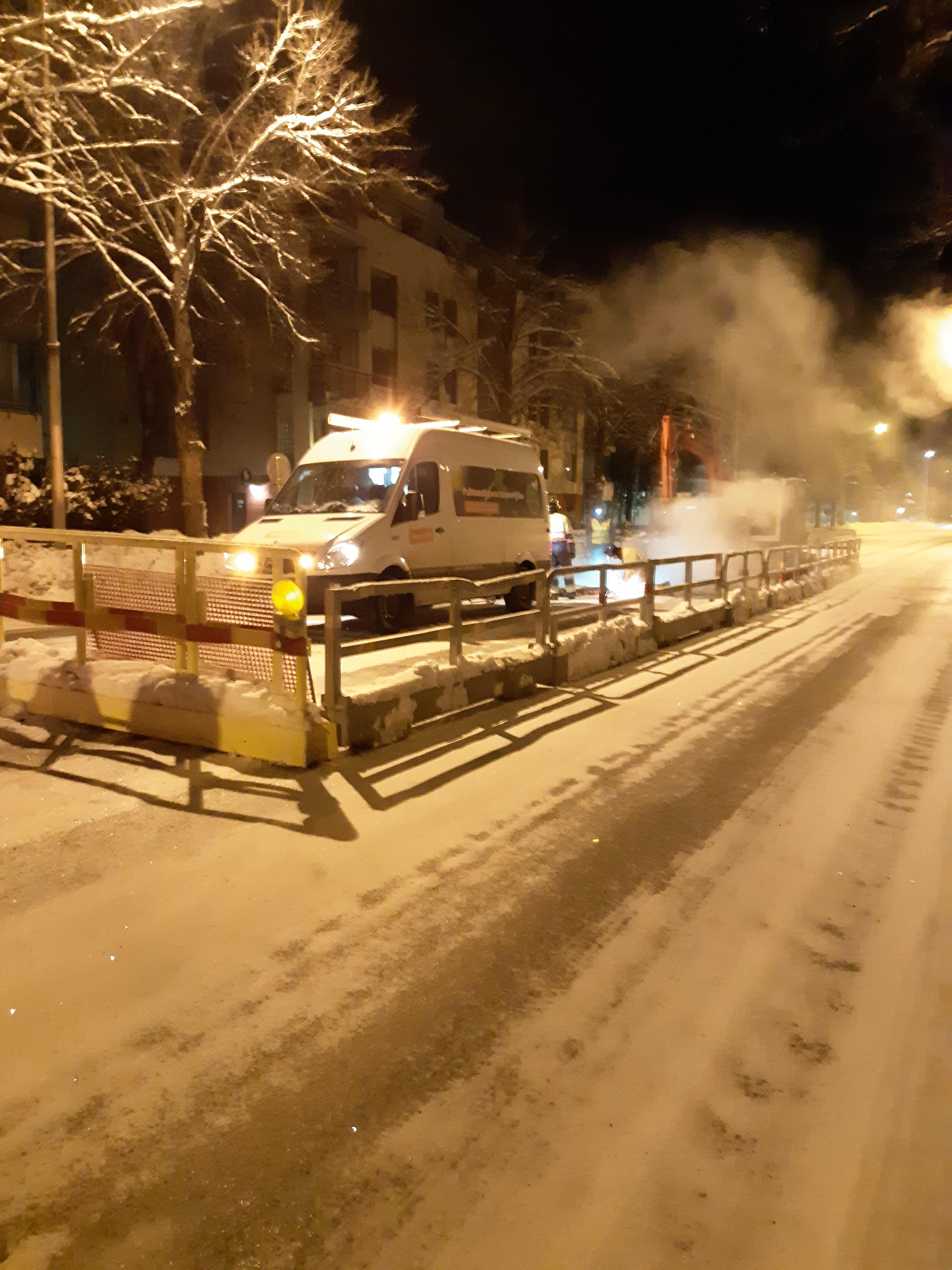 Tummaa talvitaivasta vasten nousee höyryä aidatulla alueella olevasta kaivannosta. Etualalla pakettiauto varoitusvalot päällä.