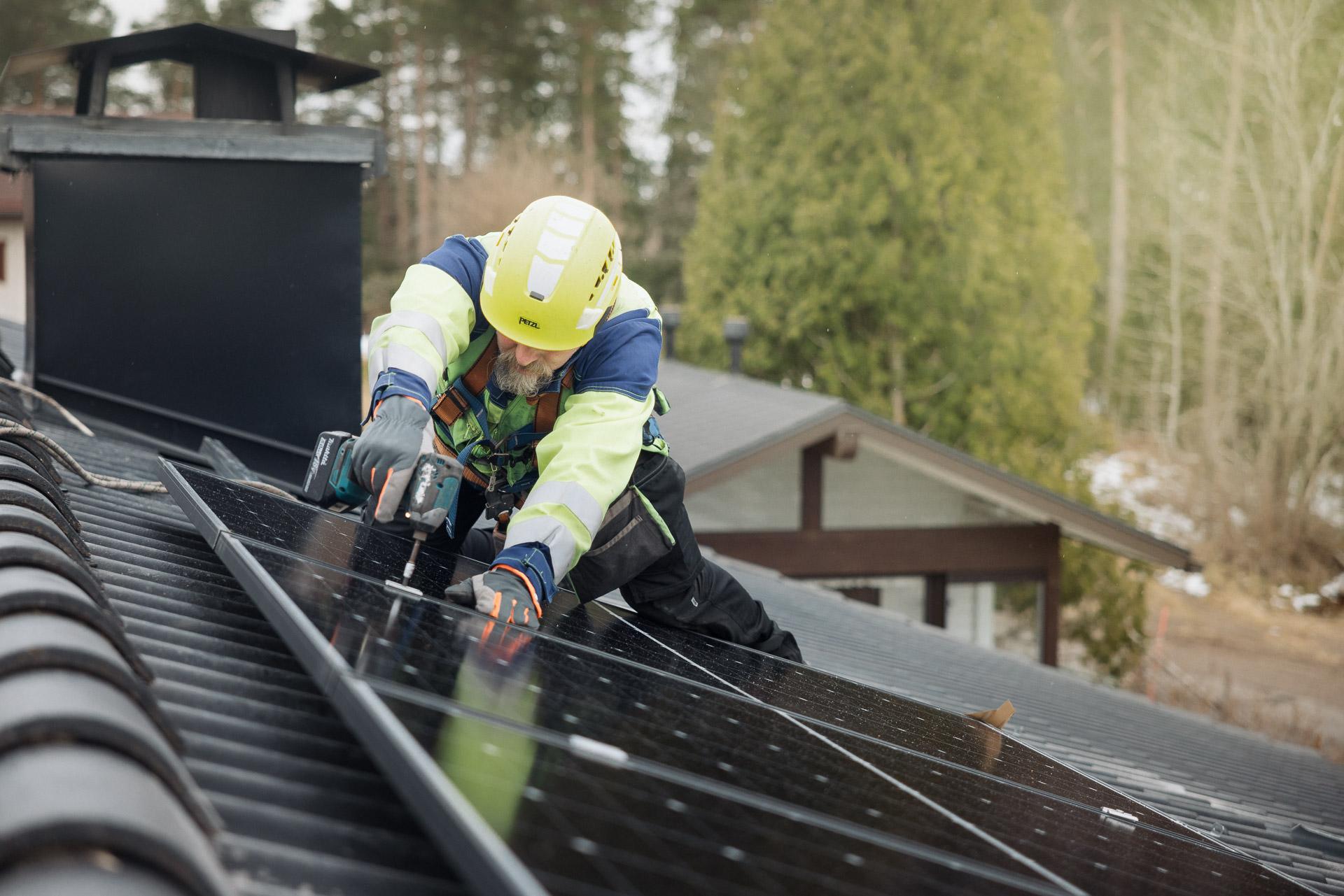 Keltaiseen kypärään ja työvaatteisiin pukeutunut mies kiinnittää aurinkopaneeleja tiilikatolle. Asentajalla on pora kädessään. Taustalla näkyy keväistä metsää ja piharakennuksen katto.